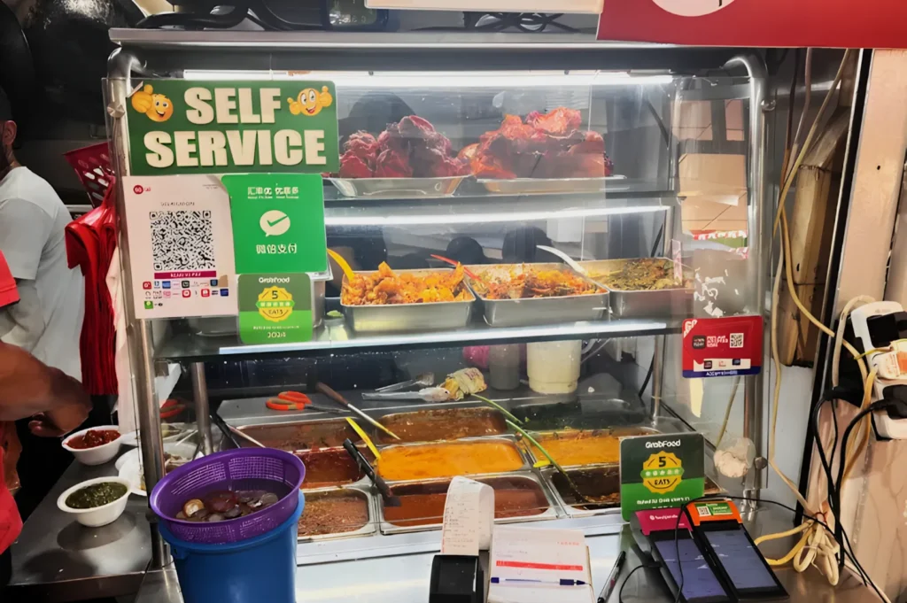 Food stall with self-service sign, displaying assorted dishes like curries and grilled items. QR codes and payment terminals are visible for easy transactions.