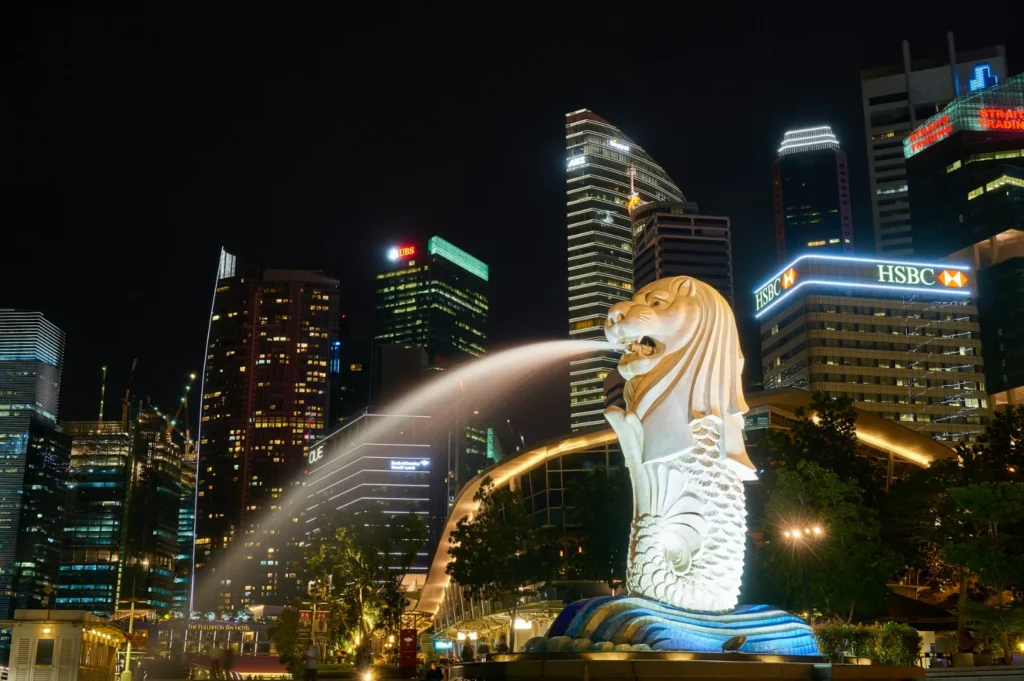 Night view of Singapore's iconic Merlion statue spouting water against a backdrop of illuminated skyscrapers, creating a vibrant urban scene.