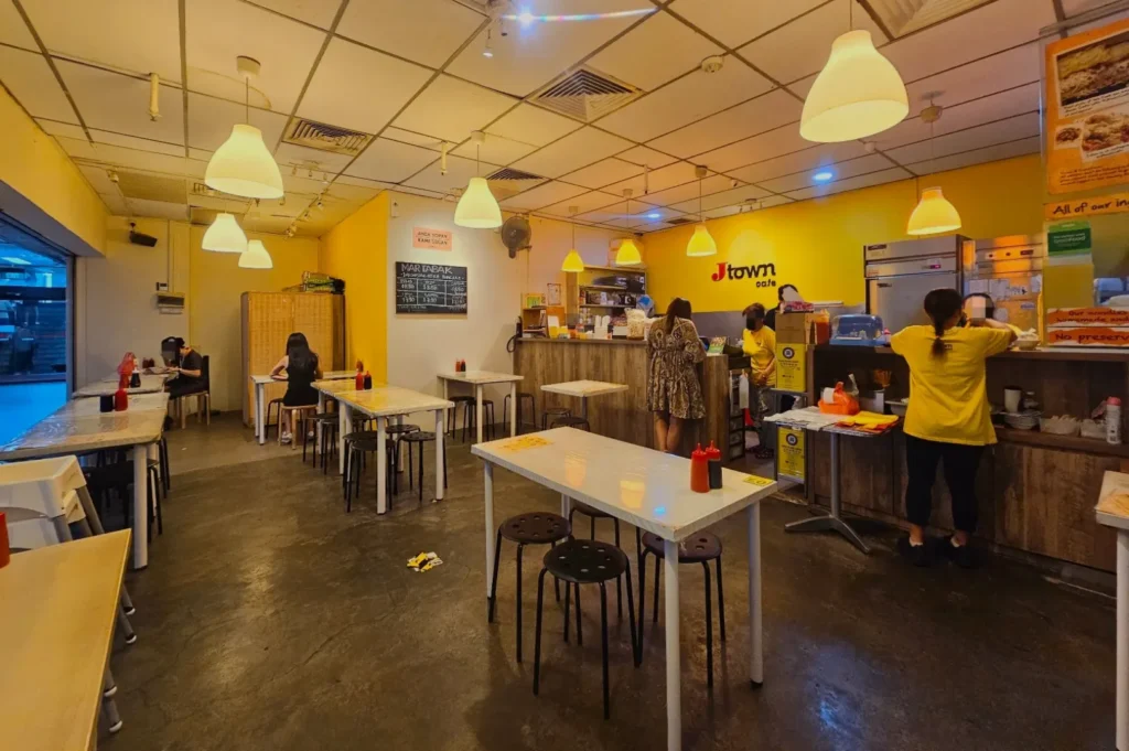 Cozy café interior with dim lighting and yellow walls. Several women in yellow shirts work behind the counter. White tables and hanging lamps fill the space, creating a warm, inviting atmosphere.