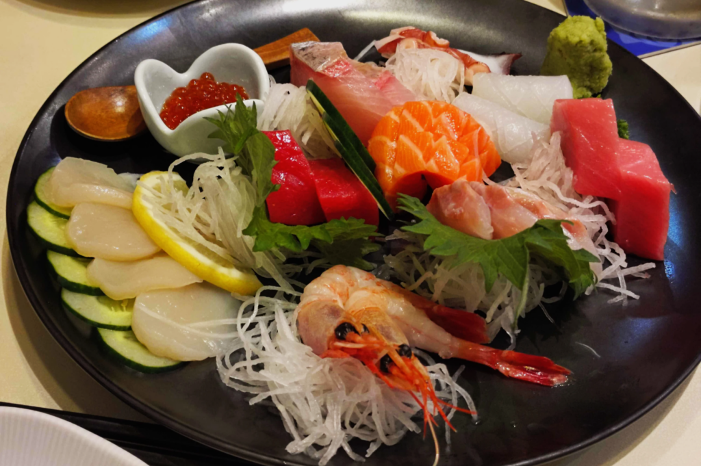 A black plate with assorted sashimi, including salmon, tuna, shrimp, and scallop. A small dish holds red fish roe. Garnished with lemon, cucumber, and wasabi.