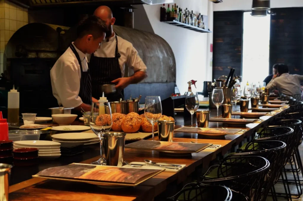 Two chefs prepare food in a restaurant kitchen. The counter is set with glasses, menus, and bread rolls. The atmosphere is busy and focused.