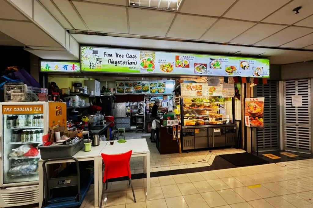 A cozy vegetarian food stall named Pine Tree Cafe. It features a bright sign, colorful meal images, a cooling tea fridge, and a welcoming red chair.