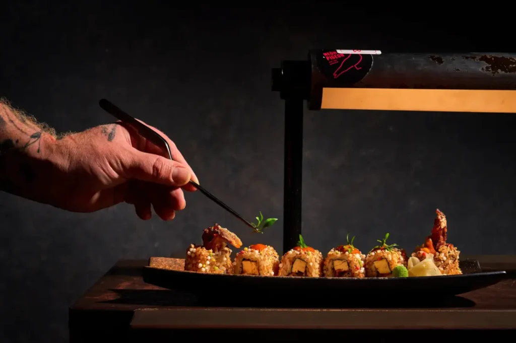 Chef's hand with tweezers garnishing sushi rolls on a dark plate, under warm light. The scene conveys precision and artistry in cuisine.