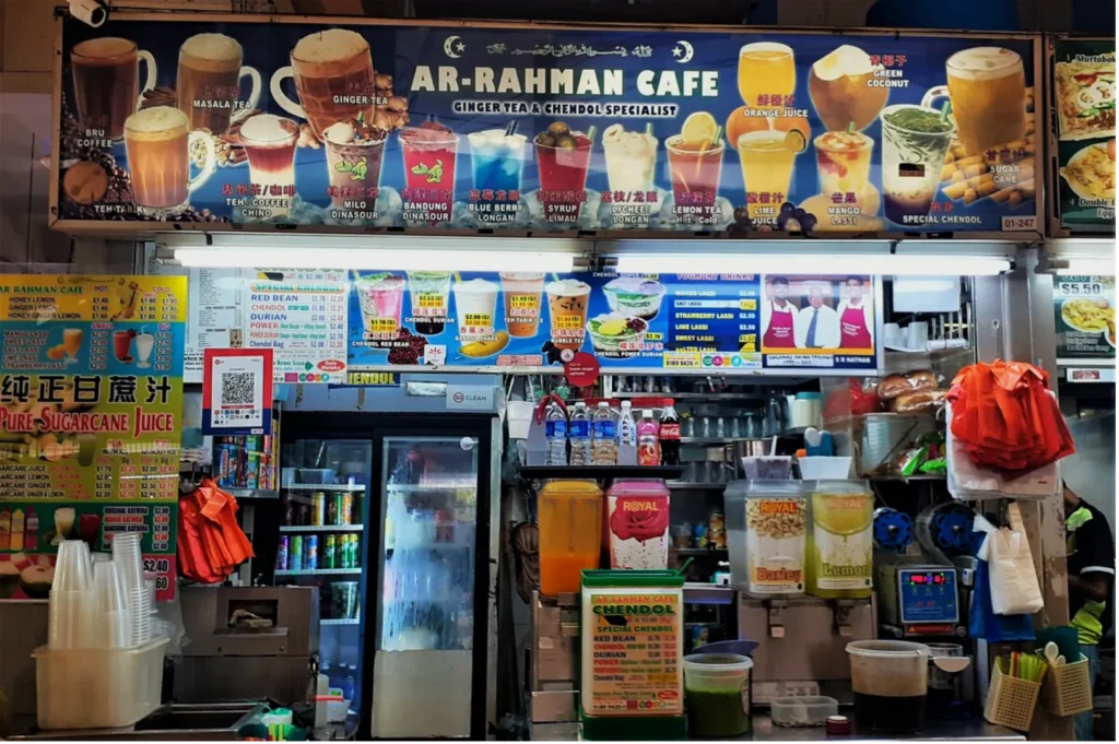 A vibrant drink stall named "Ar-Rahman Cafe" specializes in ginger tea and chendol. The menu above displays various colorful beverages, including tea, juice, and dessert drinks. The counter is filled with stacked cups, juice dispensers, and ingredients, creating a bustling and inviting atmosphere for customers.