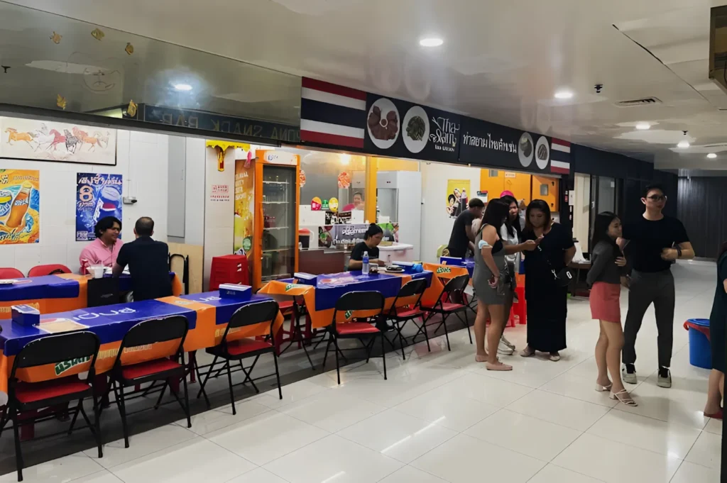 A lively Thai snack bar with an orange and blue decor. Patrons sit at tables or stand chatting. The atmosphere is casual and bustling.