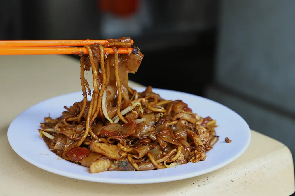 A plate of stir-fried noodles with meat and vegetables on a table. Orange chopsticks lift a portion, showcasing the dish's golden brown glaze.