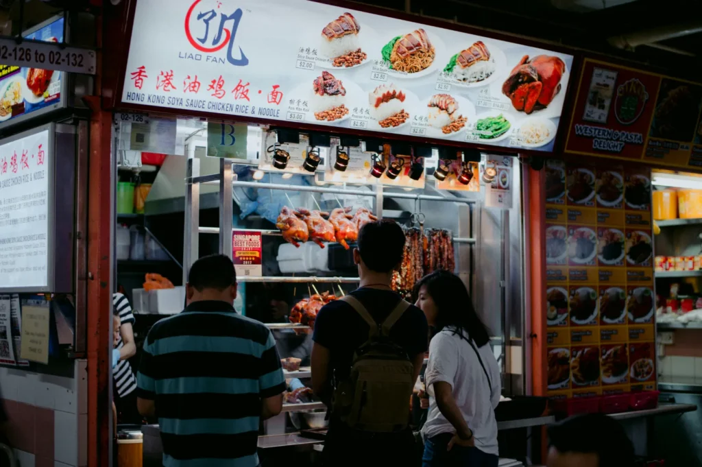 A bustling hawker stall with a bright menu board showing various Chinese dishes. Three people stand in line, eagerly awaiting their orders.