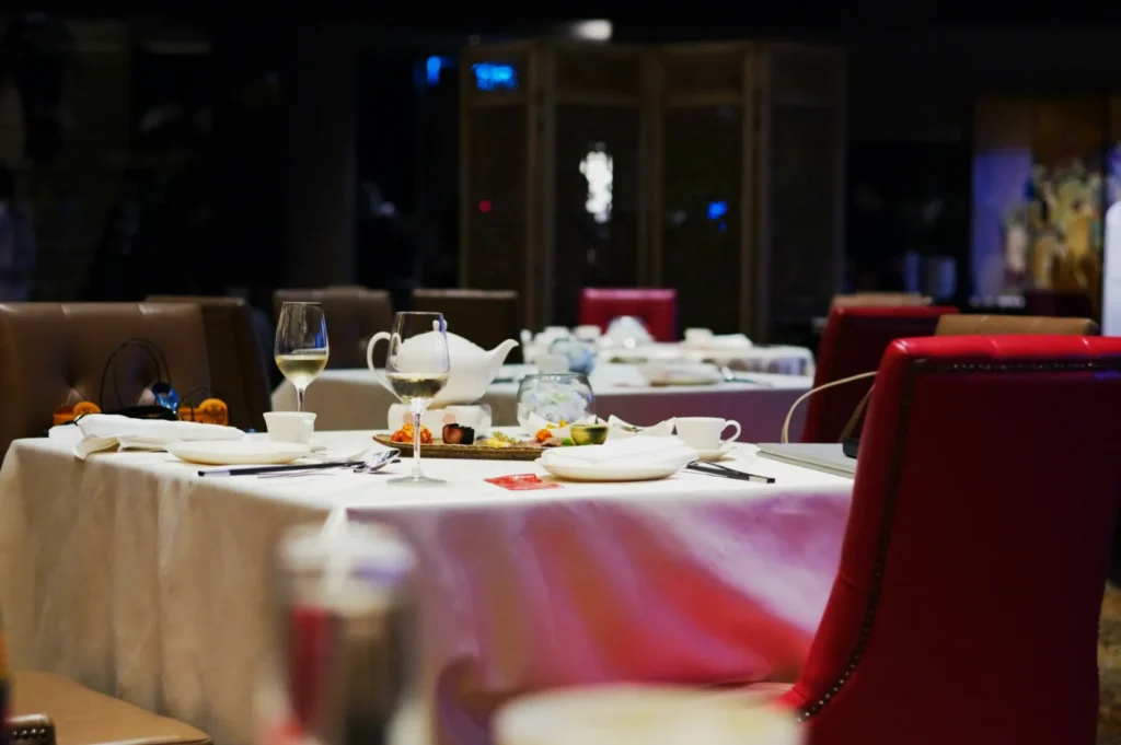 A dimly lit restaurant table set for dining with white tablecloth, red chairs, wine glasses, teapot, and plates. Background features elegant decor.