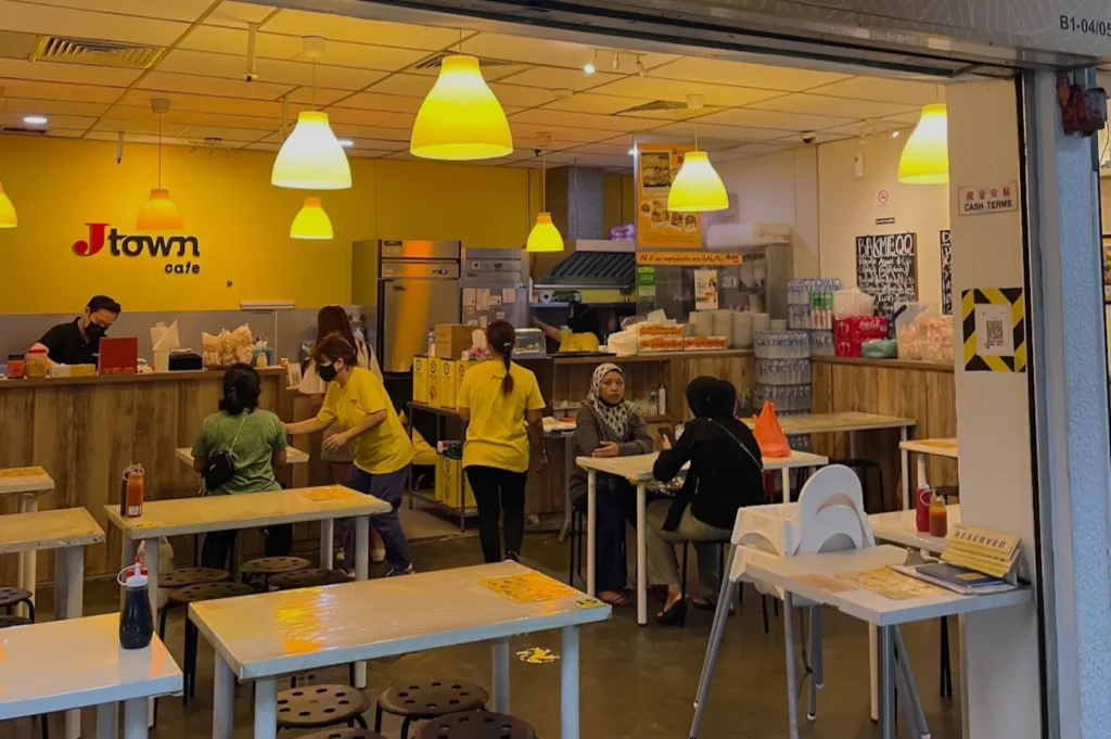 A cozy cafe with a warm yellow theme, wooden tables, and stools. Patrons are seated and chatting, while staff in yellow shirts serve food. The atmosphere is vibrant and welcoming.