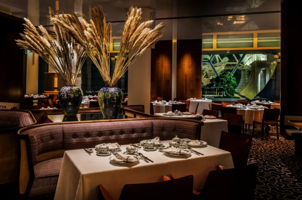Elegant restaurant interior with a cozy ambiance. Tables set with white tablecloths and fine china, two vases with tall pampas grass as centerpieces.