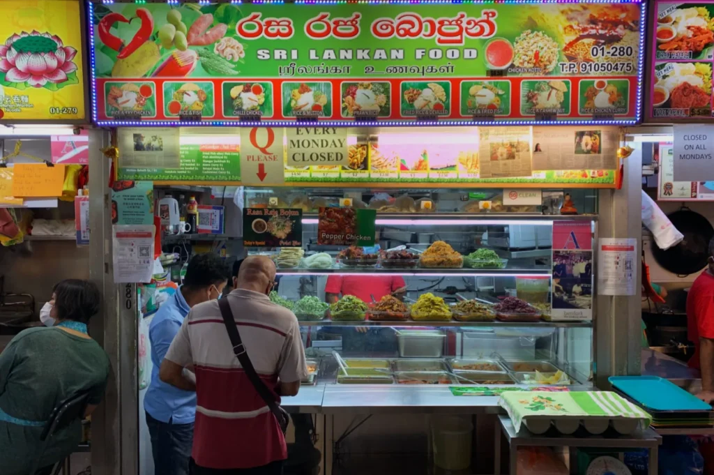 A vibrant Sri Lankan food stall with a colorful menu board overhead. Three customers wait at the counter displaying various curries. Bright, busy atmosphere.