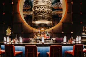 Sophisticated bar scene with a unique cylindrical chandelier featuring human faces. Red chairs and flower arrangements create an elegant, modern ambiance.