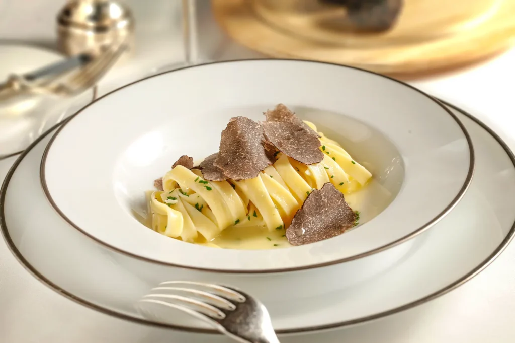 A close-up, eye-level shot captures a gourmet serving of fettuccine pasta in a light butter sauce, elegantly topped with shaved black truffles and finely chopped herbs. The pasta is presented in a wide, white rimmed bowl resting on a matching plate, set against a clean, upscale restaurant table setting with a silver fork in the foreground.