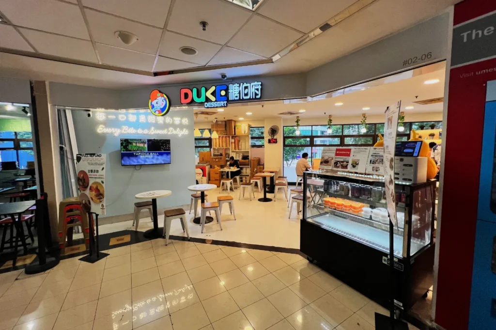 A dessert shop with a sign reading "DUKE Dessert," featuring pastel decor, small round tables, and a glass display counter with desserts. Cozy and inviting atmosphere.