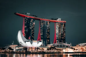 A low-angle night shot captures the iconic Marina Bay Sands complex in Singapore, featuring its three illuminated skyscrapers connected by a massive skypark illuminated with red accents. In the foreground, the distinct lotus-shaped ArtScience Museum glows bright white along the water's edge, reflecting softly on the surface.