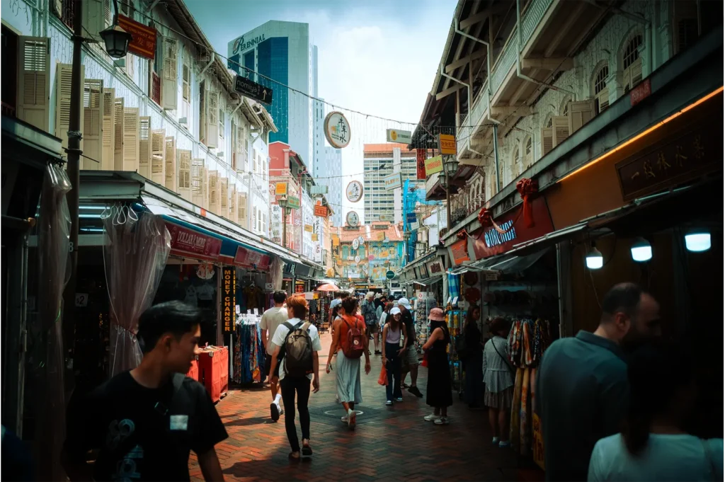 An eye-level shot captures a bustling pedestrian street in a Chinatown district, lined with historic shophouses featuring louvered shutters and various storefront awnings. Numerous pedestrians stroll along the brick-paved lane under hanging round banners, while modern high-rise skyscrapers stand in the distant background under a bright sky.