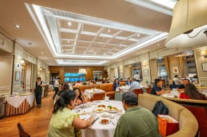 An eye-level shot captures a brightly lit, elegant Chinese restaurant dining room filled with patrons seated at round and rectangular tables with white tablecloths. The upscale interior features a large recessed coffered ceiling, wood floors, classic wall paneling, and a long tan leather booth lining the back wall.