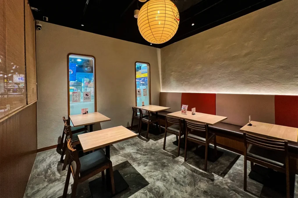 An eye-level shot captures an empty restaurant dining area featuring light wood tables paired with dark wooden chairs and a long bench with gray and red backrests. The modern space is lit by a large, spherical paper lantern hanging from the black ceiling, casting a warm glow over polished concrete floors and textured walls.