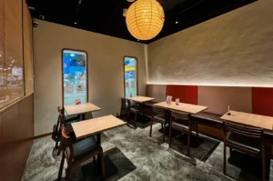 An eye-level shot captures an empty restaurant dining area featuring light wood tables paired with dark wooden chairs and a long bench with gray and red backrests. The modern space is lit by a large, spherical paper lantern hanging from the black ceiling, casting a warm glow over polished concrete floors and textured walls.