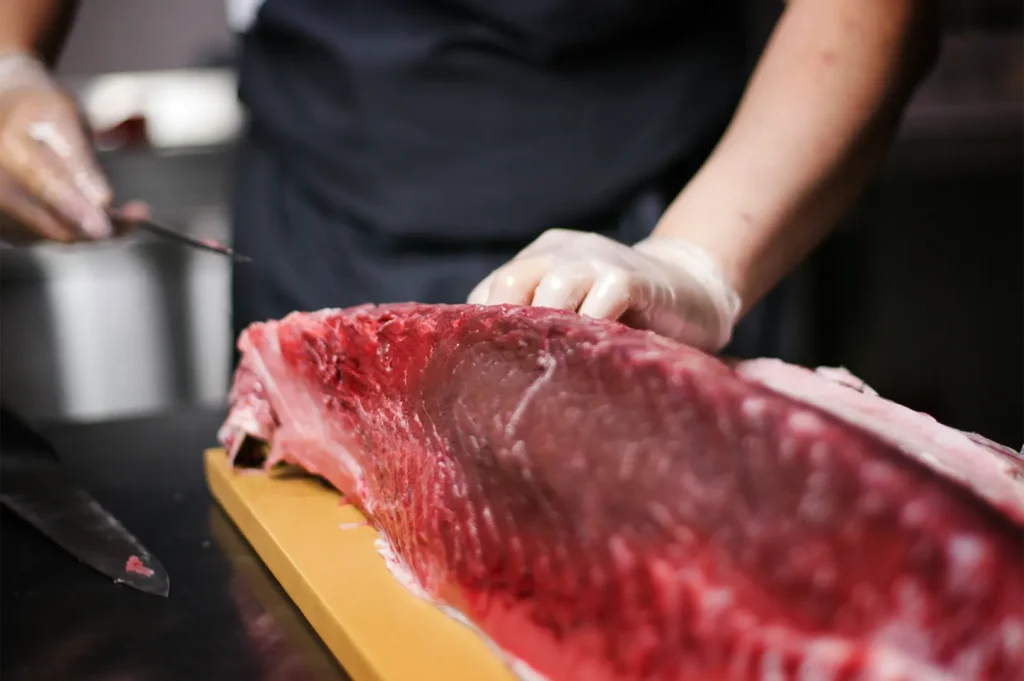 A close-up shot captures a chef wearing clear plastic gloves butchering a massive, deep red loin of fresh tuna on a wooden cutting board. The chef holds a long, sharp knife in one hand while stabilizing the premium cut of fish with the other.