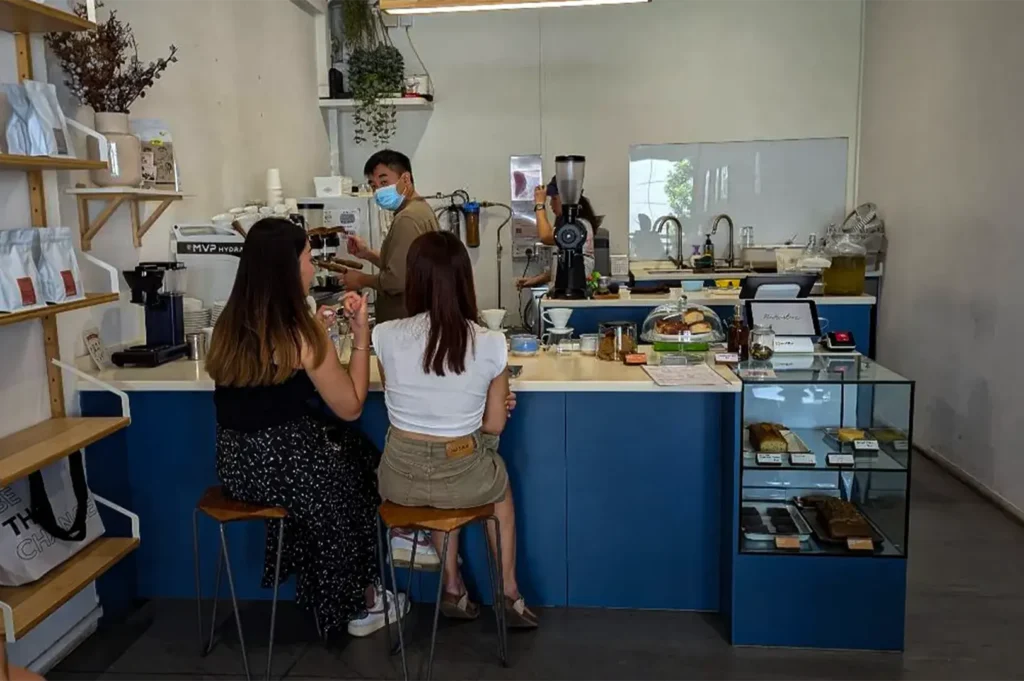 An eye-level, medium shot captures the bright, minimalist interior of a modern coffee shop where customers and baristas interact at a blue-faced service counter. In the foreground, two women sit on wooden bar stools with slim metal legs; the woman on the left wears a black top with a patterned maxi skirt, while the woman on the right wears a white crop top and khaki shorts. Behind the counter, a male barista wearing a medical face mask works near coffee-grinding equipment, while another staff member is visible near a washing station in the background. The counter is organized with a point-of-sale tablet, a glass display case showcasing pastries, and a set of wooden shelves on the far left wall neatly stocked with white bags of coffee beans.