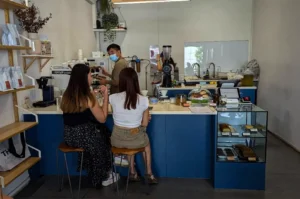 An eye-level, medium shot captures the bright, minimalist interior of a modern coffee shop where customers and baristas interact at a blue-faced service counter. In the foreground, two women sit on wooden bar stools with slim metal legs; the woman on the left wears a black top with a patterned maxi skirt, while the woman on the right wears a white crop top and khaki shorts. Behind the counter, a male barista wearing a medical face mask works near coffee-grinding equipment, while another staff member is visible near a washing station in the background. The counter is organized with a point-of-sale tablet, a glass display case showcasing pastries, and a set of wooden shelves on the far left wall neatly stocked with white bags of coffee beans.