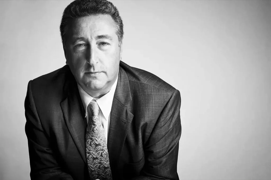 A close-up, black-and-white studio portrait captures a man with short, textured hair looking directly forward with a calm, neutral expression. He is dressed in a dark, subtly patterned suit jacket worn over a white collared shirt and a light-colored paisley tie. Leaning slightly forward, the man is positioned on the left side of the frame, while the bright, solid white background on the right creates a clean and minimalist composition.