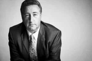 A close-up, black-and-white studio portrait captures a man with short, textured hair looking directly forward with a calm, neutral expression. He is dressed in a dark, subtly patterned suit jacket worn over a white collared shirt and a light-colored paisley tie. Leaning slightly forward, the man is positioned on the left side of the frame, while the bright, solid white background on the right creates a clean and minimalist composition.