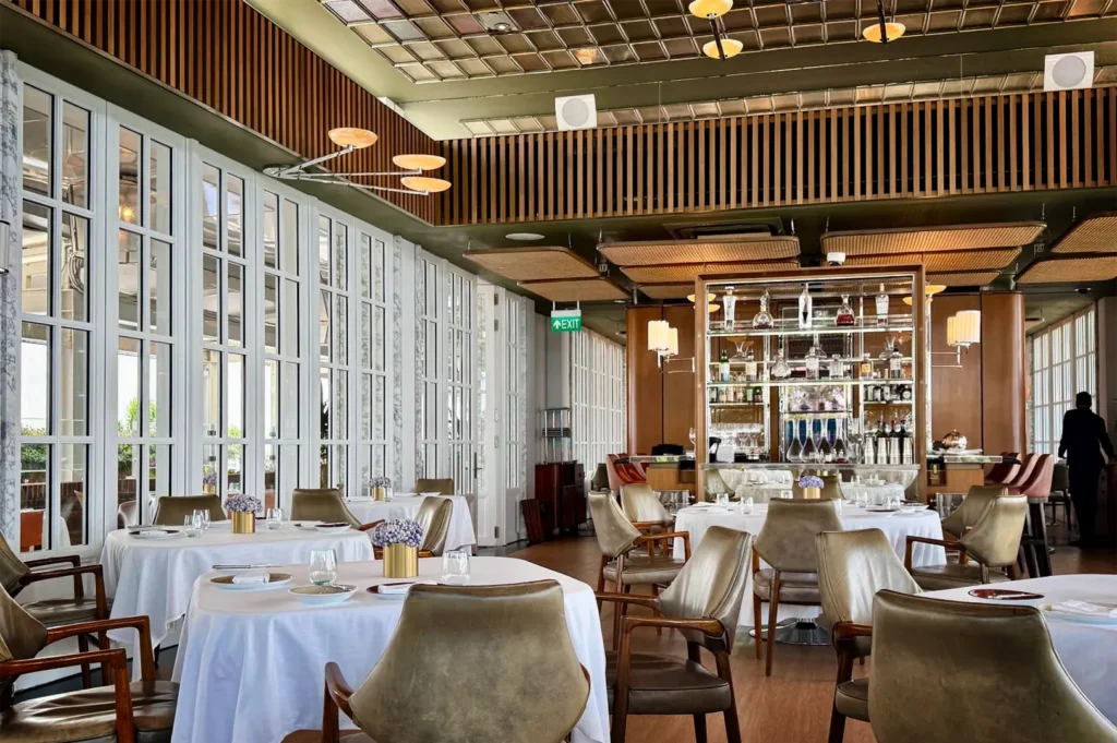 A wide, eye-level shot captures the bright and sophisticated dining room of a high-end restaurant, featuring crisp white-clothed tables paired with elegant leather and wood armchairs over smooth wooden floors. On the left, a long wall composed of floor-to-ceiling white multi-paned windows floods the space with natural light. In the background, a central freestanding bar display case with mirrored backing and glass shelves showcases bottles and decanters under ambient lighting, while a sleek wood-slat accent band wraps around the upper perimeter below a grid-patterned ceiling with unique modern light fixtures.
