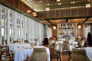A wide, eye-level shot captures the bright and sophisticated dining room of a high-end restaurant, featuring crisp white-clothed tables paired with elegant leather and wood armchairs over smooth wooden floors. On the left, a long wall composed of floor-to-ceiling white multi-paned windows floods the space with natural light. In the background, a central freestanding bar display case with mirrored backing and glass shelves showcases bottles and decanters under ambient lighting, while a sleek wood-slat accent band wraps around the upper perimeter below a grid-patterned ceiling with unique modern light fixtures.