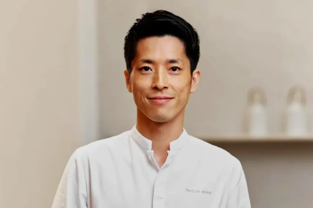 A man in a white chef's coat with a slight smile stands against a neutral background. The setting conveys professionalism and calm.