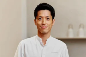 A man in a white chef's coat with a slight smile stands against a neutral background. The setting conveys professionalism and calm.