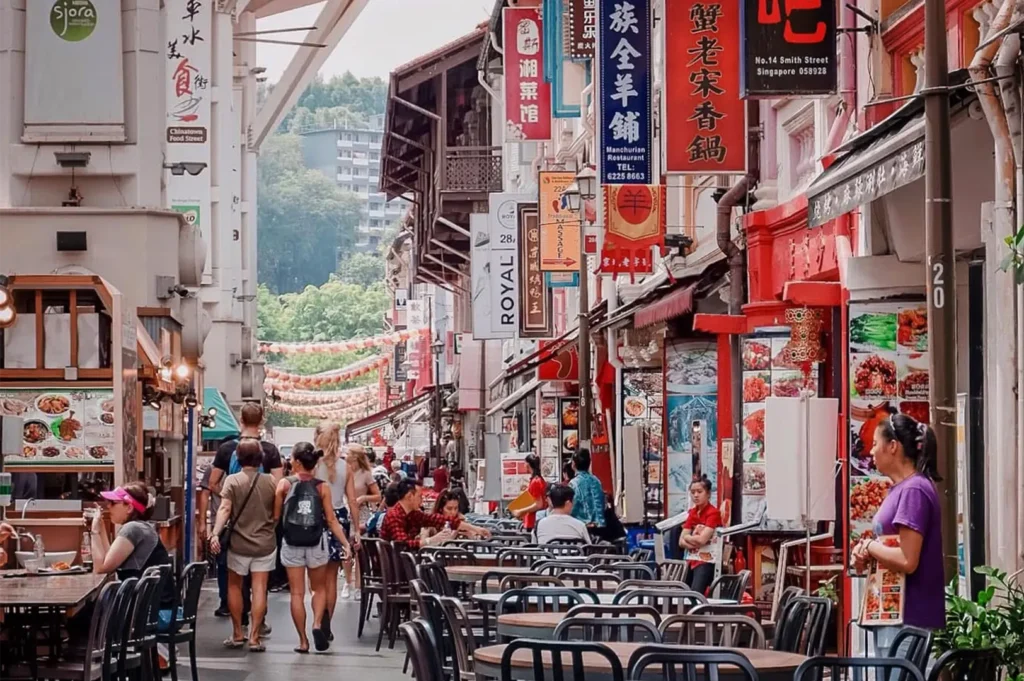An eye-level shot captures a bustling pedestrian food street lined with colorful shophouses and vibrant vertical Chinese and English signage. Pedestrians walk past outdoor dining tables and food stalls, with decorative paper lanterns hanging across the street in the background.