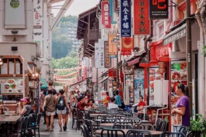 An eye-level shot captures a bustling pedestrian food street lined with colorful shophouses and vibrant vertical Chinese and English signage. Pedestrians walk past outdoor dining tables and food stalls, with decorative paper lanterns hanging across the street in the background.