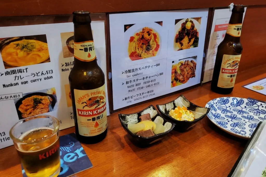 A cozy restaurant scene with Kirin beer bottles and a glass on a wooden table, alongside two dishes of pickled appetizers. Menu images and blue-patterned plates are visible in the background. The atmosphere is casual and inviting.