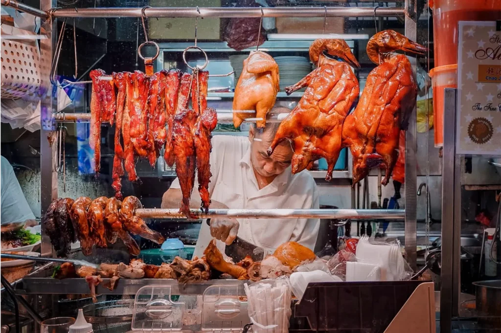 A close-up shot through a glass display window captures a food stall vendor in a white shirt chopping meat with a cleaver at his workstation. Hanging from metal racks above him is an enticing array of traditional roasted meats, including glistening strips of barbecue pork, a whole poached chicken, and golden-brown roasted ducks.