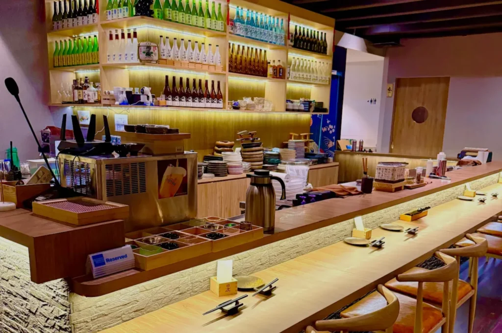Cozy sushi bar with wooden counter and high chairs. Shelves display colorful sake bottles, creating a warm, inviting ambiance. Plates and chopsticks are neatly arranged.