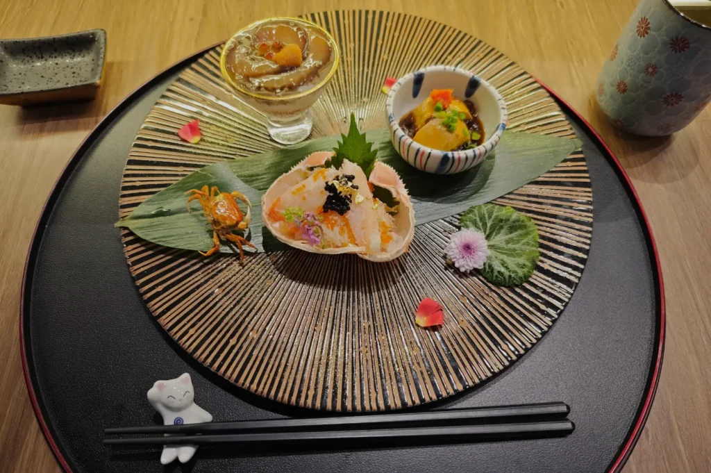 Elegant Japanese dish arrangement on a decorative plate. Features seafood in a shell, a glass of jelly, a small bowl, garnished with petals and leaves. A serene, artistic presentation.