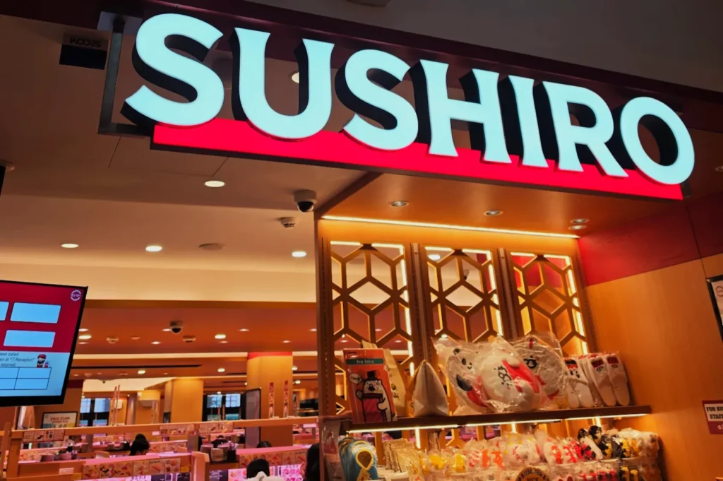 Brightly lit "Sushiro" sign in a stylish sushi restaurant with decorative wood panels and plush toys on display, creating a lively, inviting ambiance.