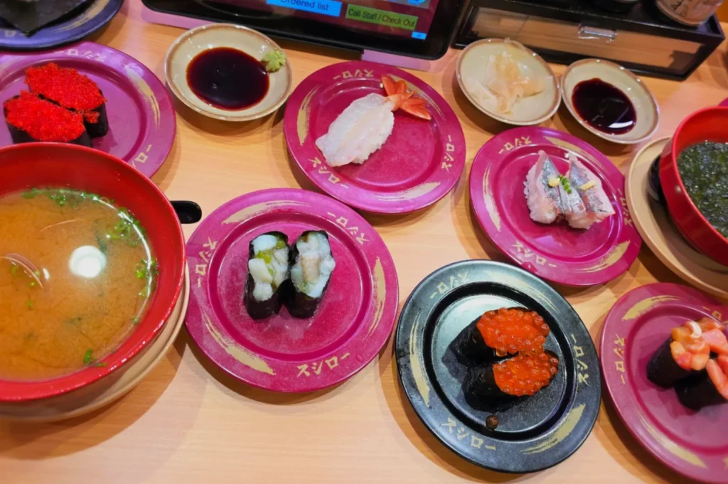 A colorful spread of sushi on plates with various toppings like shrimp, roe, and fish, accompanied by bowls of miso soup and dipping sauces.