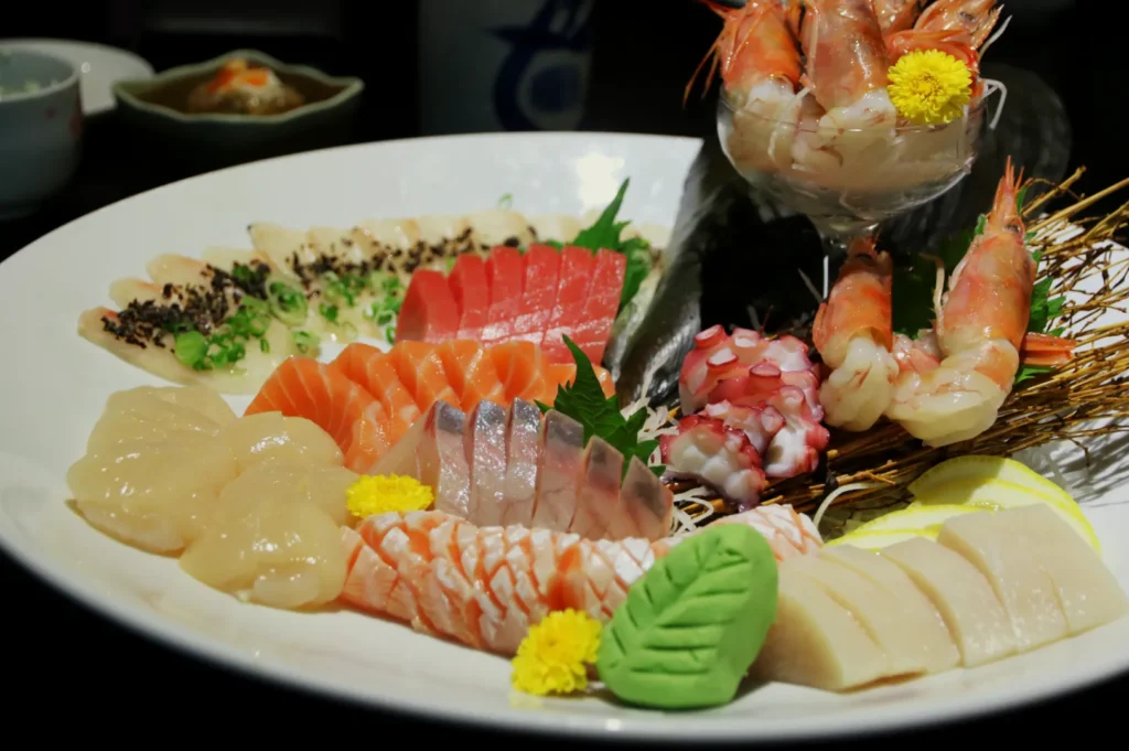 A beautifully arranged platter of assorted sashimi, featuring salmon, tuna, shrimp, and scallops, garnished with green leaves and yellow flowers.