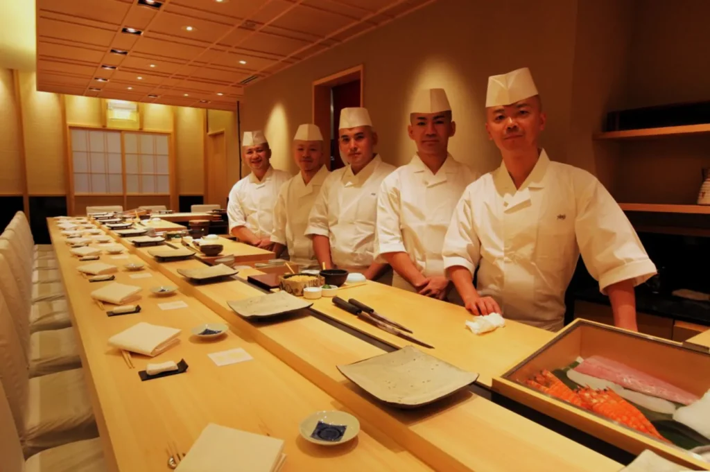 Five sushi chefs in white uniforms and hats stand proudly behind a wooden sushi bar, lined with plates and chopsticks, in a warmly lit restaurant.