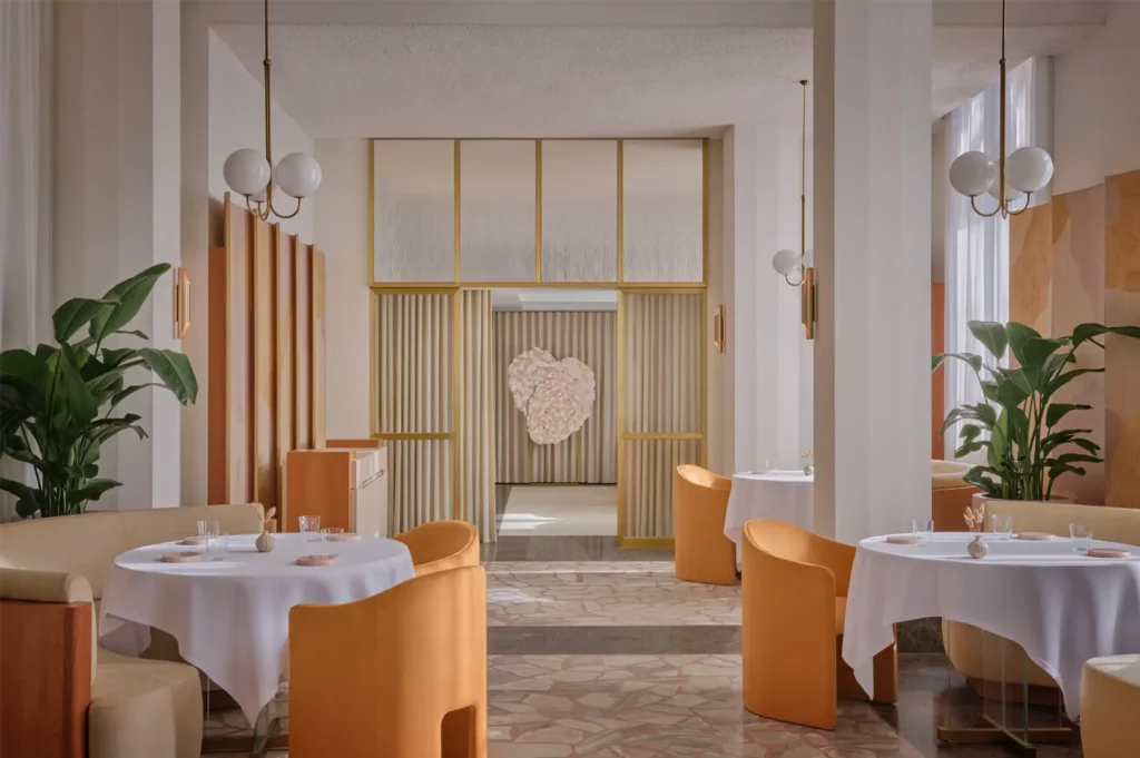An eye-level shot captures an elegant, modern restaurant interior featuring round tables with white tablecloths and plush, yellow-orange barrel chairs. The bright space is accented by terrazzo tile flooring, large potted green plants, brass light fixtures, and a heart-shaped floral installation visible through a golden-trimmed partition wall in the background.