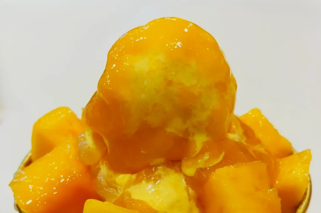 Close-up of a dessert featuring a scoop of mango ice cream topped with mango chunks and syrup on a white surface, conveying a fresh, tropical vibe.