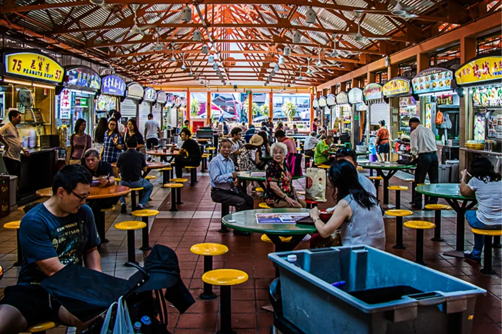 A wide eye-level shot captures a bustling indoor hawker center filled with diners seated at round tables under a high, orange-trussed metal roof. Lined with food stalls featuring colorful signage on both sides, the lively market hall shows people eating, chatting, and walking through the tiled aisles.