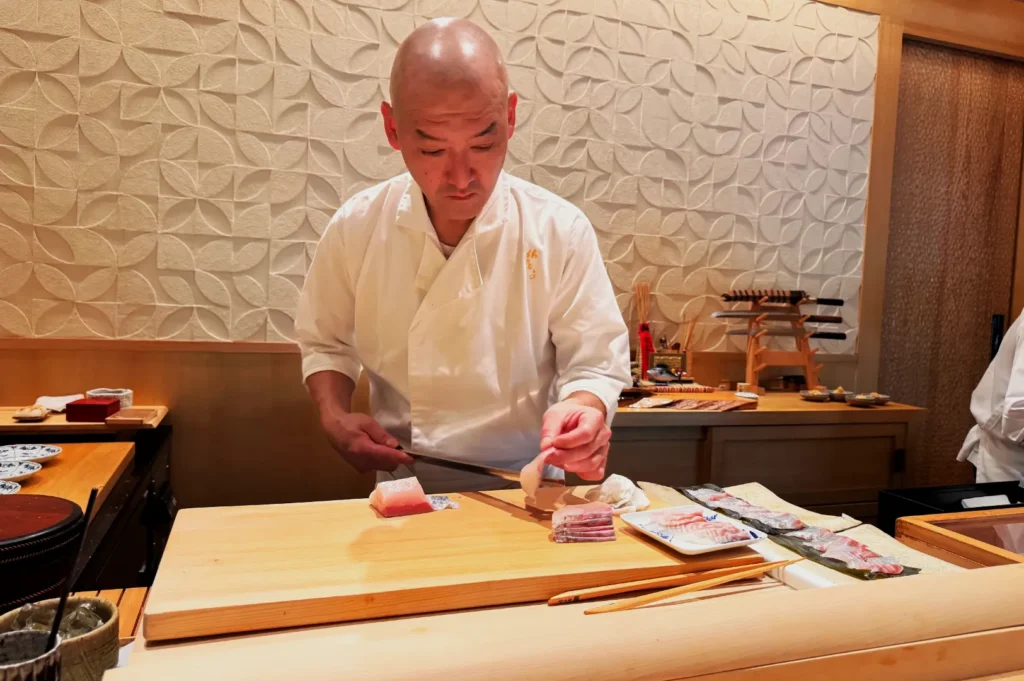 A chef in a white uniform skillfully slices fish at a sushi counter. The backdrop features a textured wall, creating a serene and focused atmosphere.