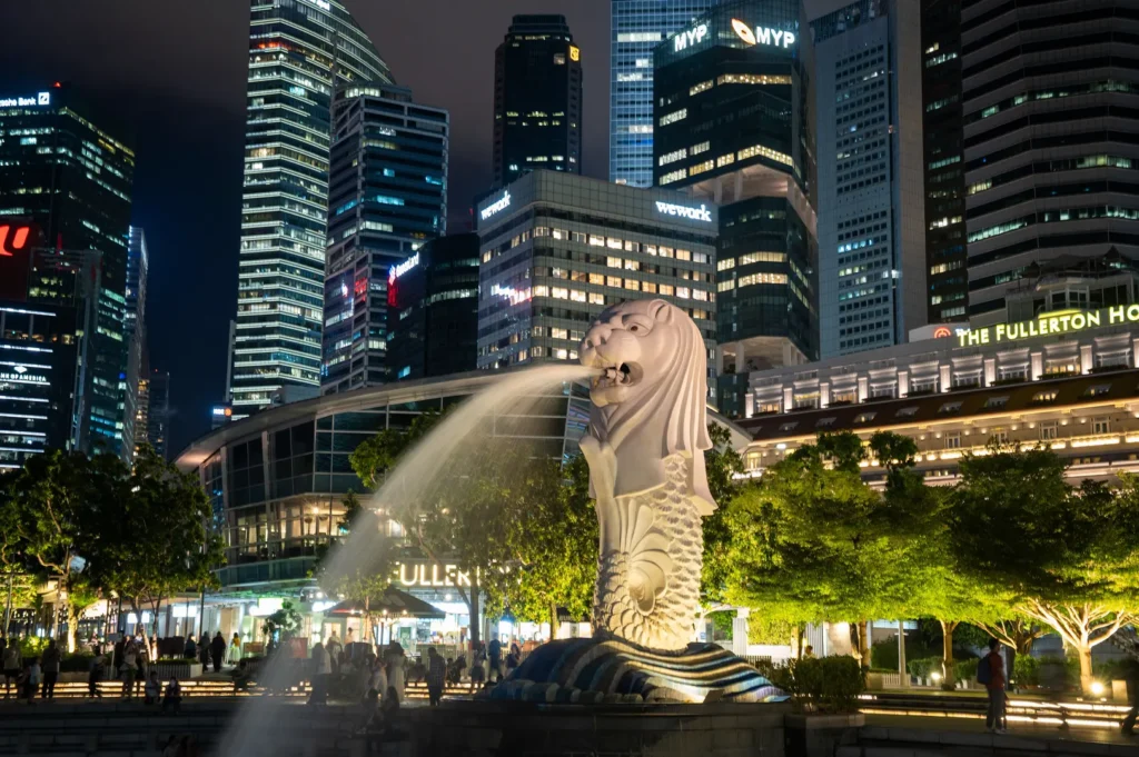 An eye-level shot captures the iconic Merlion statue illuminated at night as it spouts a continuous stream of water, surrounded by brightly lit green trees. In the background, a cluster of modern skyscrapers towers over the historic Fullerton Hotel, lighting up the dark night sky.