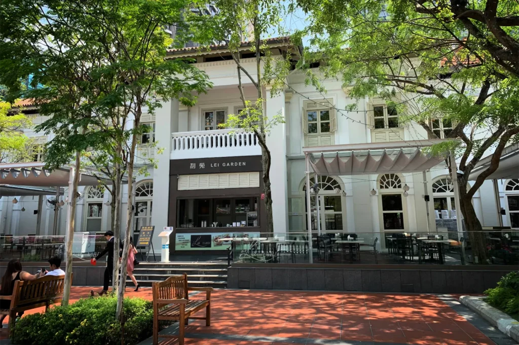 An eye-level shot captures the exterior of "Lei Garden" restaurant, housed in a historic white colonial-style building with arched windows and a prominent second-floor balcony. In the foreground, an outdoor dining patio with glass railings sits adjacent to a brick-paved walkway shaded by lush green trees.