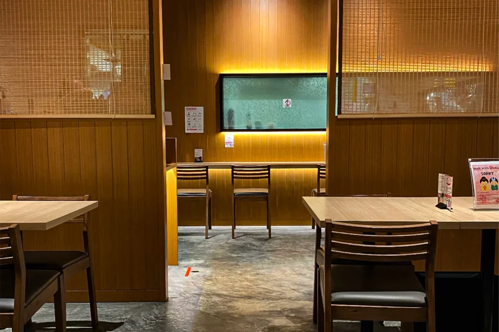 An eye-level shot captures an empty restaurant interior featuring wooden dining tables in the foreground and a counter seating area framed by partition walls in the background. The space is illuminated by warm, ambient lighting accentuating the vertical wood paneling and a back-lit rectangular window or display case.
