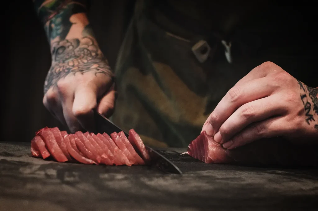 A close-up shot captures a chef with tattooed hands slicing a fresh block of raw tuna on a dark cutting board. Using a sharp knife, the chef has neatly lined up thin sashimi slices on the left while stabilizing the remaining portion of the fish with their right hand.